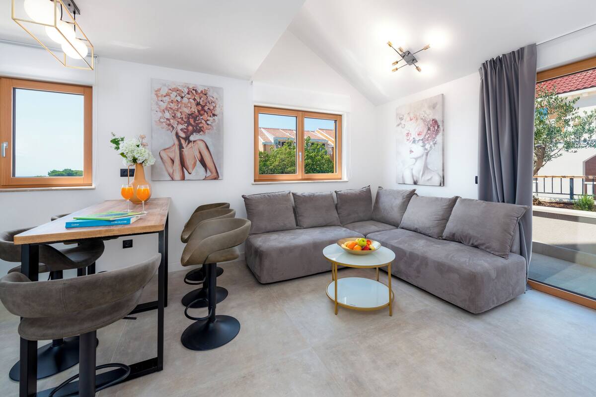 A modern living room with a grey sofa, a round coffee table with fruit, a wooden desk with books, and two chairs. There are two windows with wooden frames and a view of houses outside.
