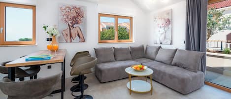Un salon moderne avec un canapé d’angle, une table à manger avec des chaises, une petite table ronde sur laquelle se trouve un saladier rempli de fruits, et des œuvres d’art accrochées aux murs.