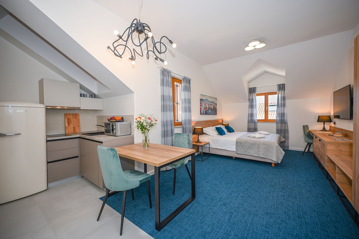 A modern hotel room with a kitchenette, a dining area, a bed, and a desk.