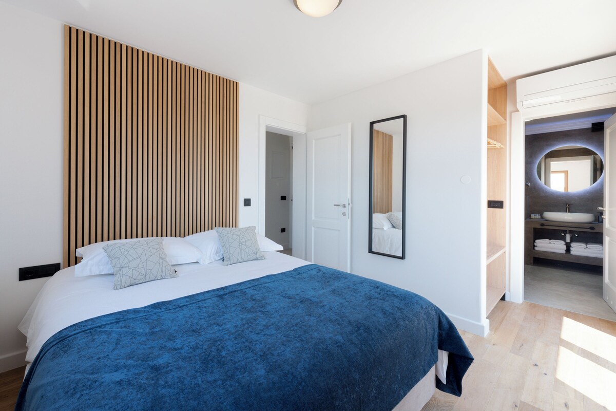Un dormitorio con cama, pared de listones de madera, espejo y baño visible a través de una puerta abierta.