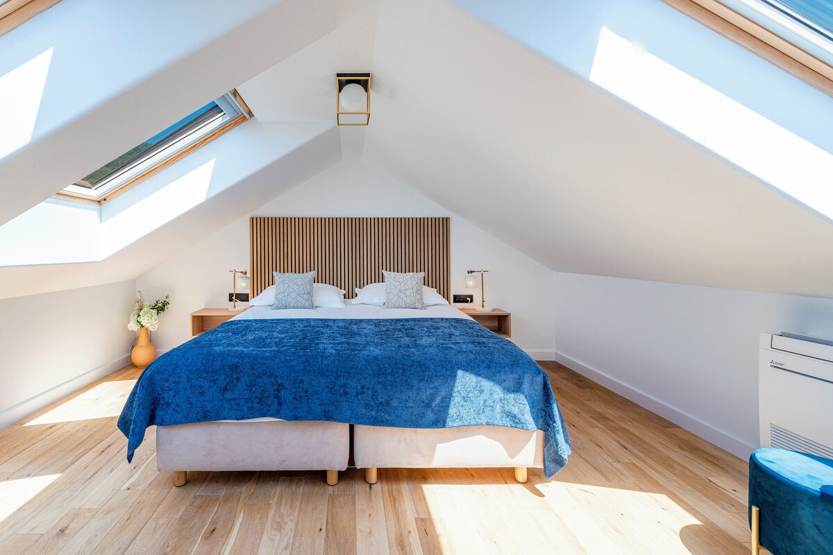 A bedroom with a sloped ceiling, a skylight, a bed with a blue bedspread, wooden headboard, bedside tables, and a nightstand with a lamp.