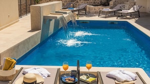 A pool area with a water feature, lounge chairs, and a table with drinks and snacks. - Casa Laurea (Dubrovnik)