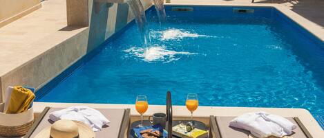 A pool area with a water feature, lounge chairs, and a table with drinks and snacks.