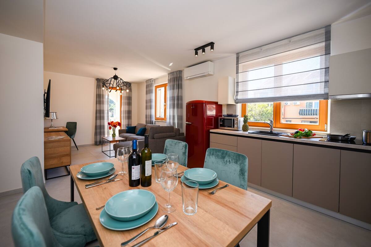 A modern kitchen with a red refrigerator, a dining area with teal chairs and a wooden table set for four, and a living area with a sofa and a coffee table.