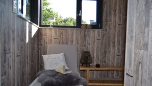 A bedroom with a wooden wall, a bed with a white pillow, a small table with a candle, and a window with a view of trees. - Tasteful vacation loft for 6 people in Emsland with sauna and outdoor pool (Emsbüren)