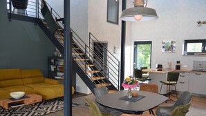 A modern living room with a yellow sofa, a dining table with chairs, a staircase, and a kitchen area. - Tasteful vacation loft for 6 people in Emsland with sauna and outdoor pool (Emsbüren)