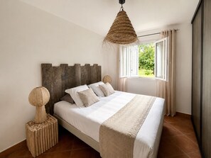 A bedroom with a wooden headboard, a bed with white bedding, a woven hanging light, and a view of greenery through the window.