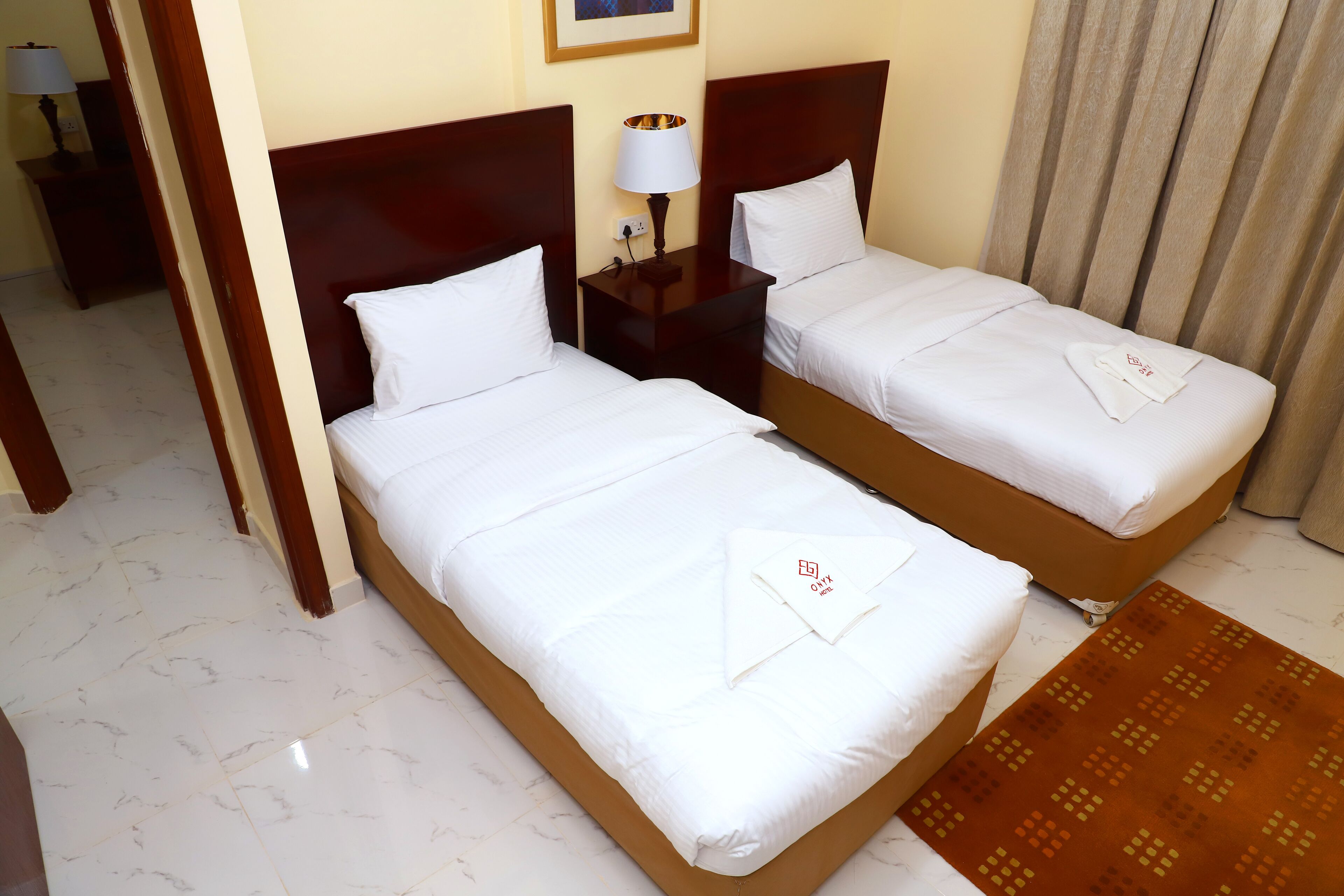Two neatly made beds with white linens and towels, a small bedside table with a lamp, and a patterned rug on a tiled floor.