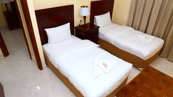 Two neatly made beds with white linens and towels, a small bedside table with a lamp, and a patterned rug on a tiled floor.
