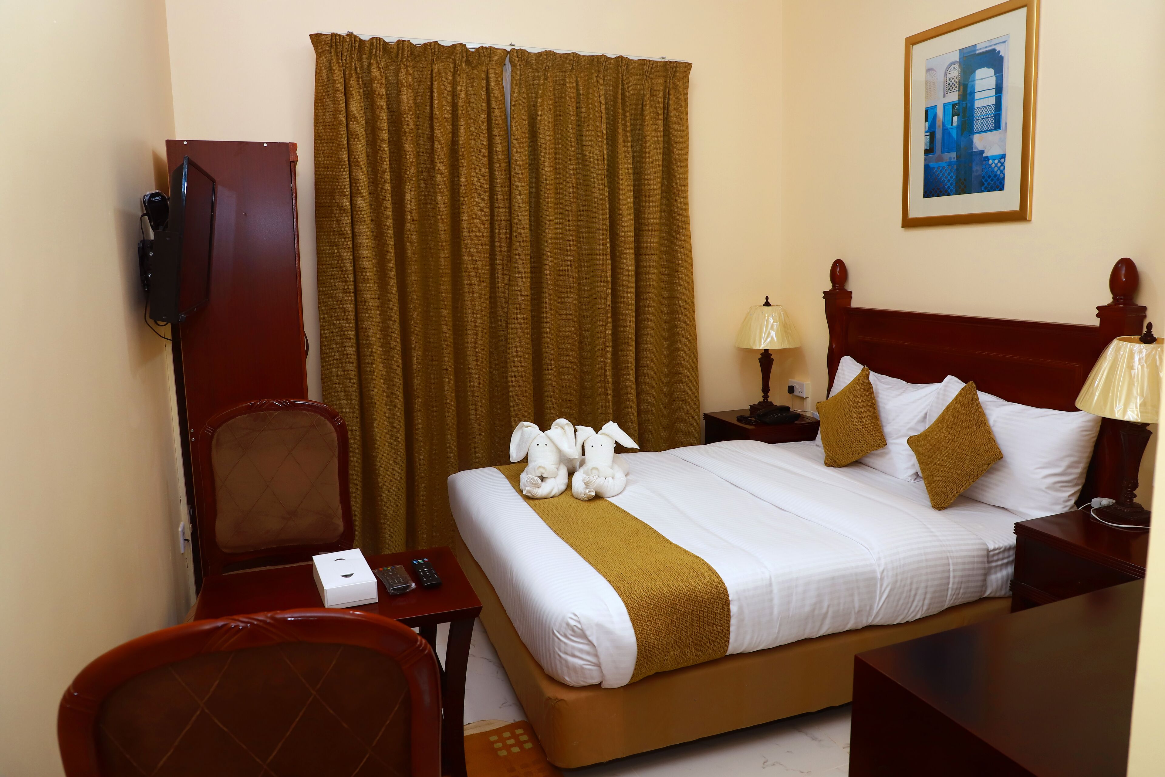 A hotel room with a bed, a desk, a chair, a TV, a lamp, and a painting on the wall.