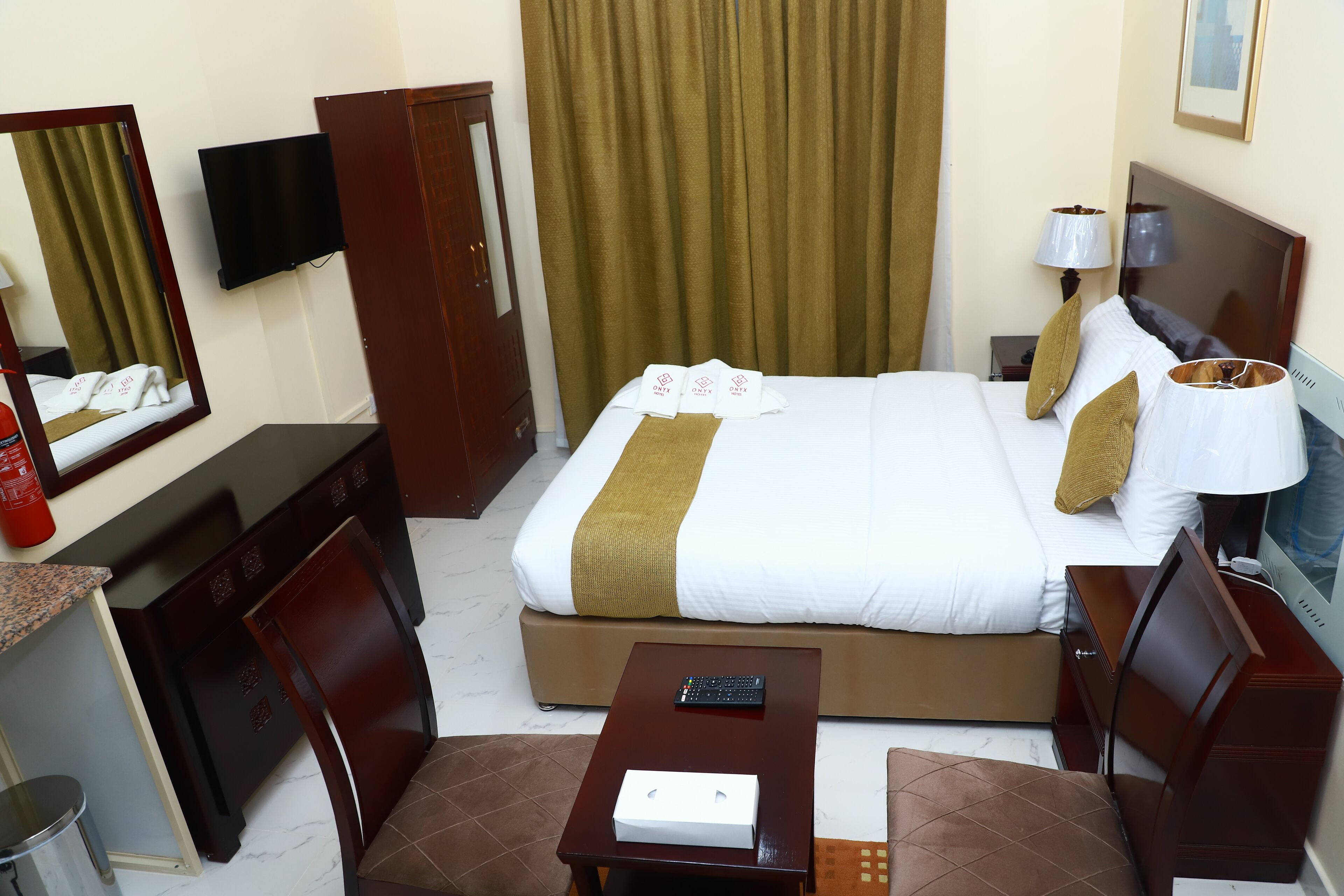 A hotel room with a bed, a desk with a remote control, a chair, a TV on the wall, and a window with curtains.