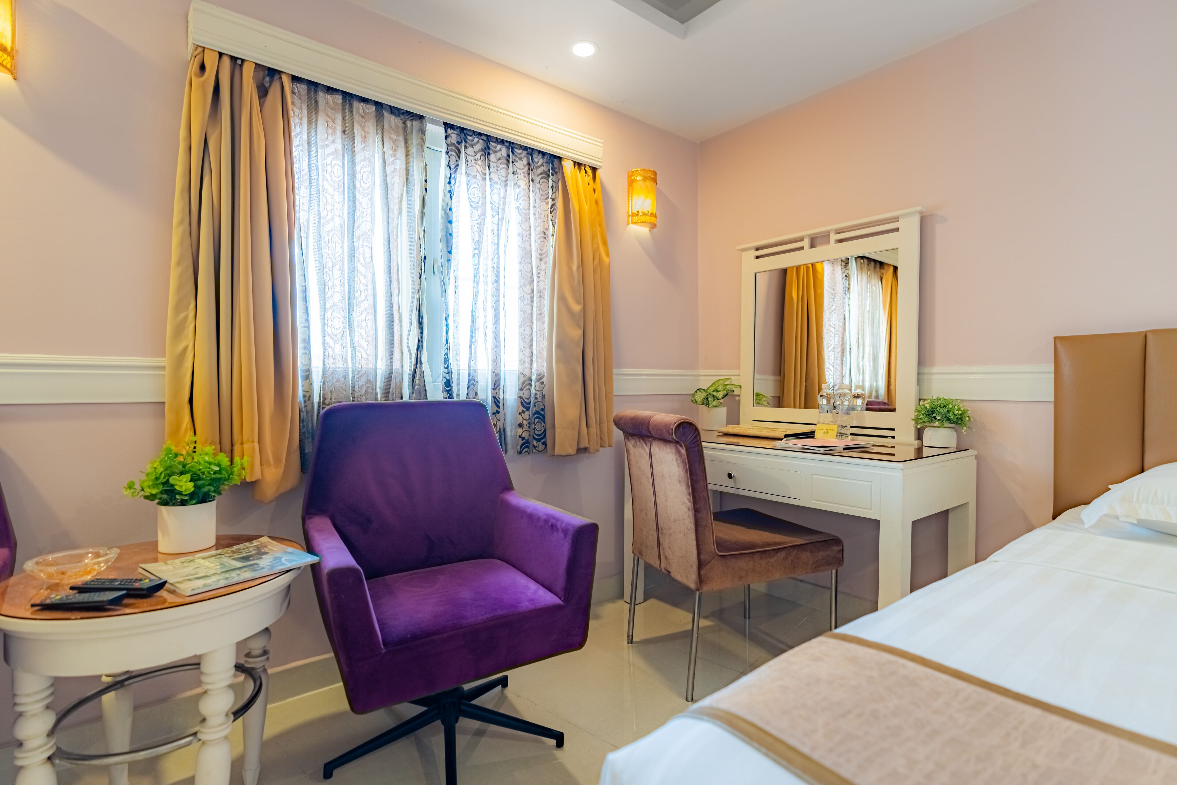 A hotel room with a bed, a desk with a chair, a small table with a plant, and a window with curtains.
