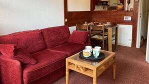 Un espacio de sala acogedor con un sofá rojo, una mesa de centro de madera con tazas, una mesa de comedor lista para cuatro personas y un cuadro de un carruaje tirado por caballos en la pared.