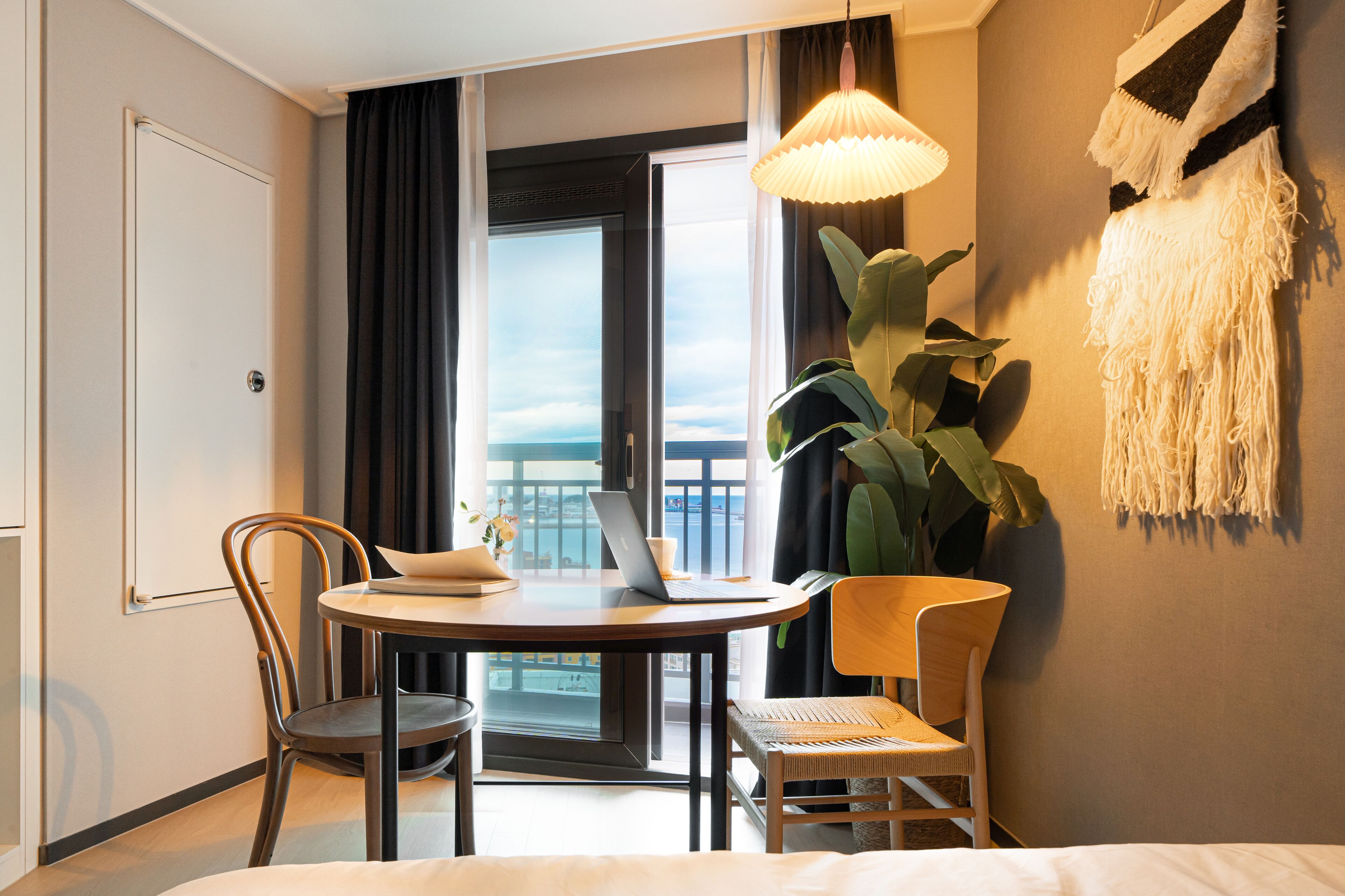 A hotel room with a bed, a desk with a laptop, two chairs, a large plant, and a view of the sea through a window.