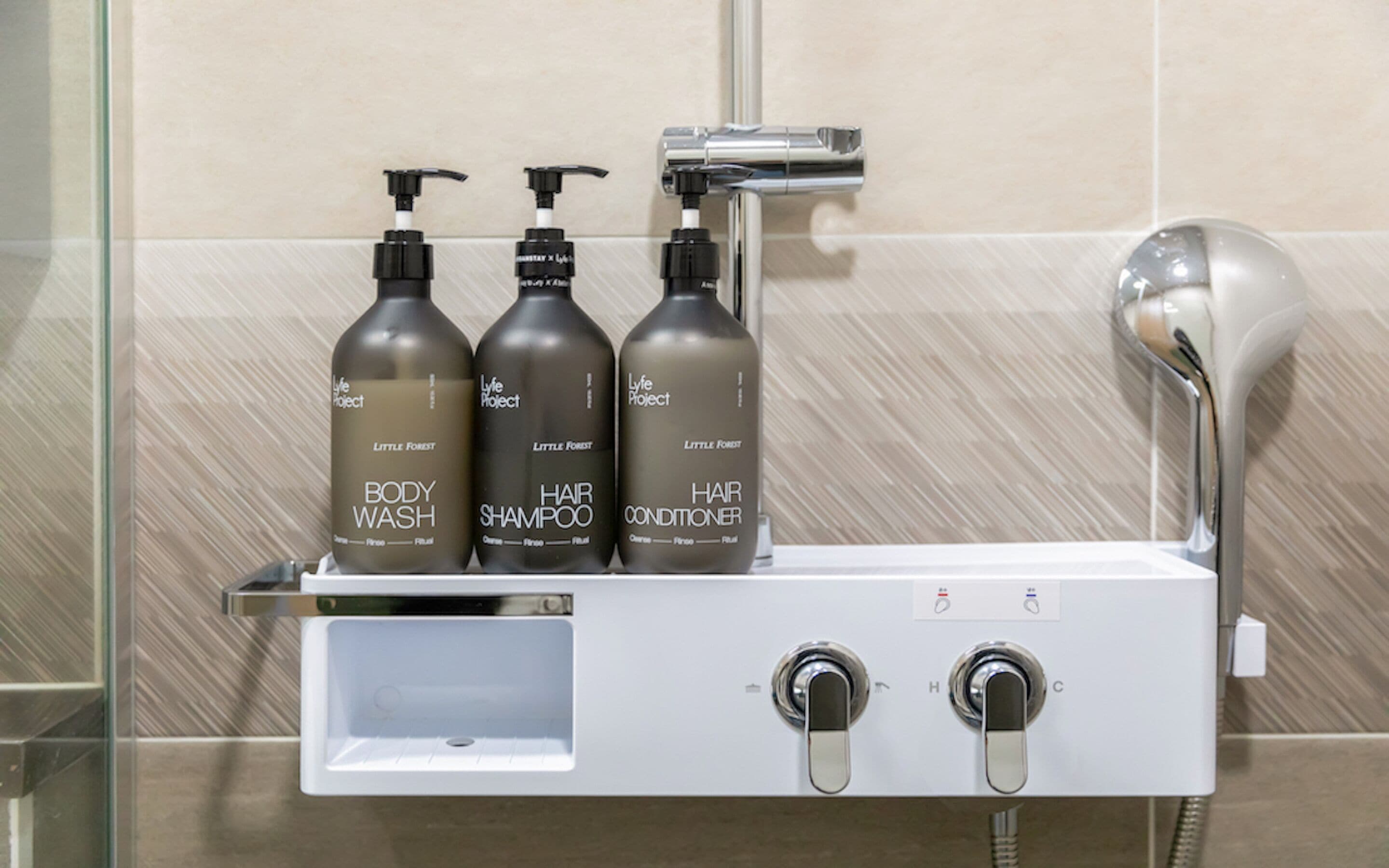 A bathroom with a shower, a shelf holding three bottles of body wash, hair shampoo, and hair conditioner. There are two knobs on the wall, likely for controlling water temperature and flow.