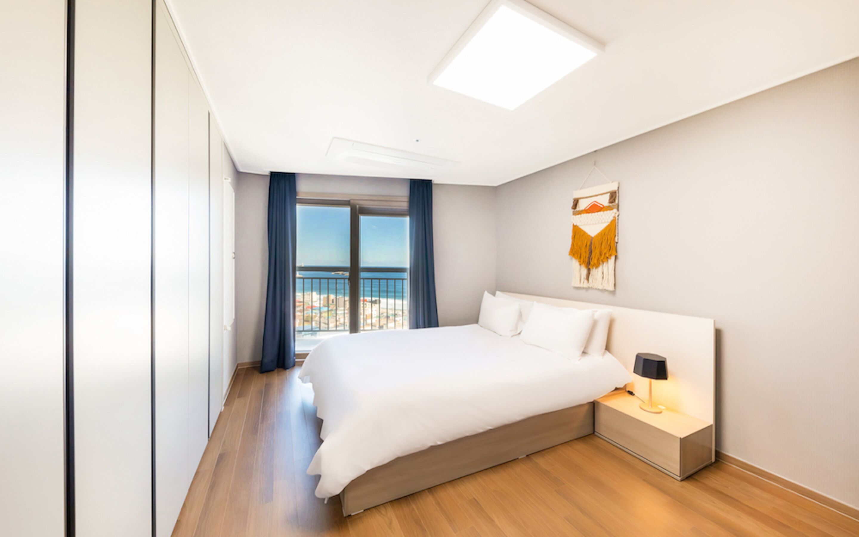A modern hotel room with a large bed, a nightstand with a lamp, a view of the sea through a balcony, and a ceiling light.