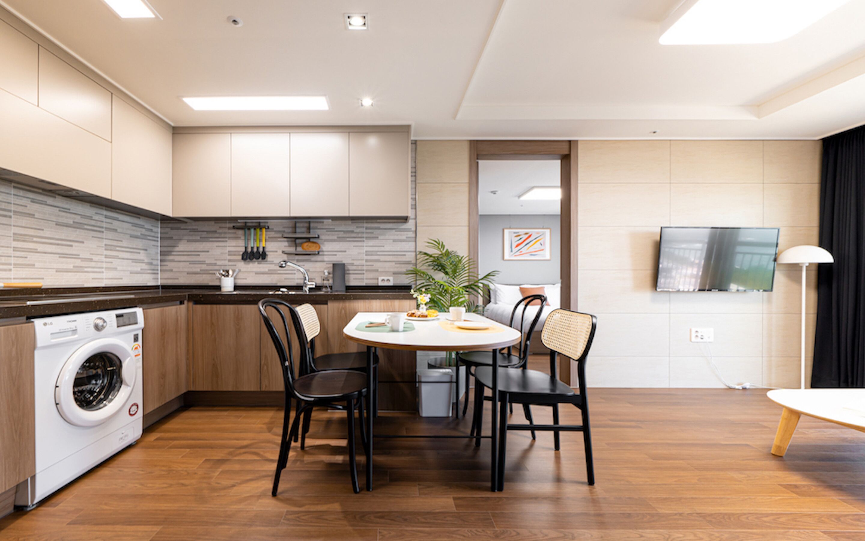 A modern kitchen with a built-in washing machine, a dining area with a round table and chairs, and a flat-screen TV mounted on the wall.