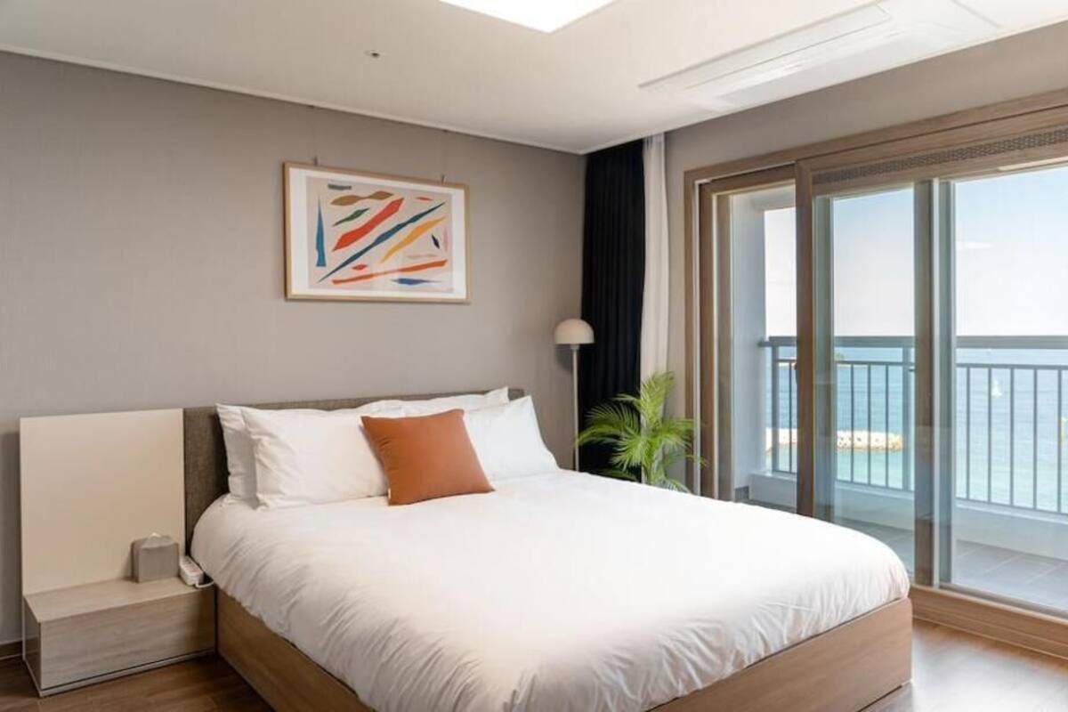 Ein modernes Hotelzimmer mit einem großen Bett, einem Nachttisch, einer Lampe, Blick auf das Meer durch eine Schiebetür und einem gerahmten abstrakten Gemälde an der Wand.