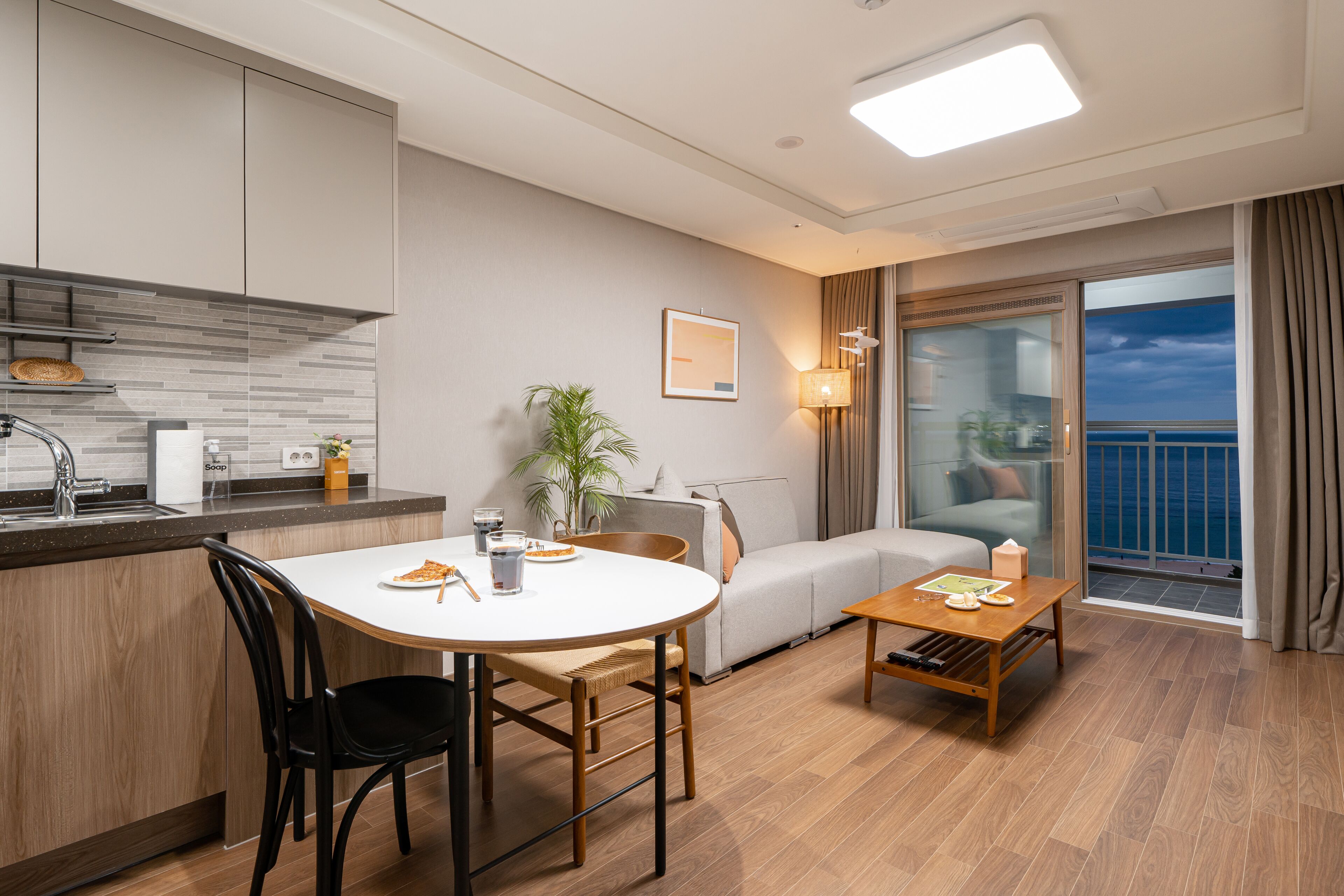 A modern hotel room with a kitchen area, a dining table, a sofa, and a balcony with a view of the sea.