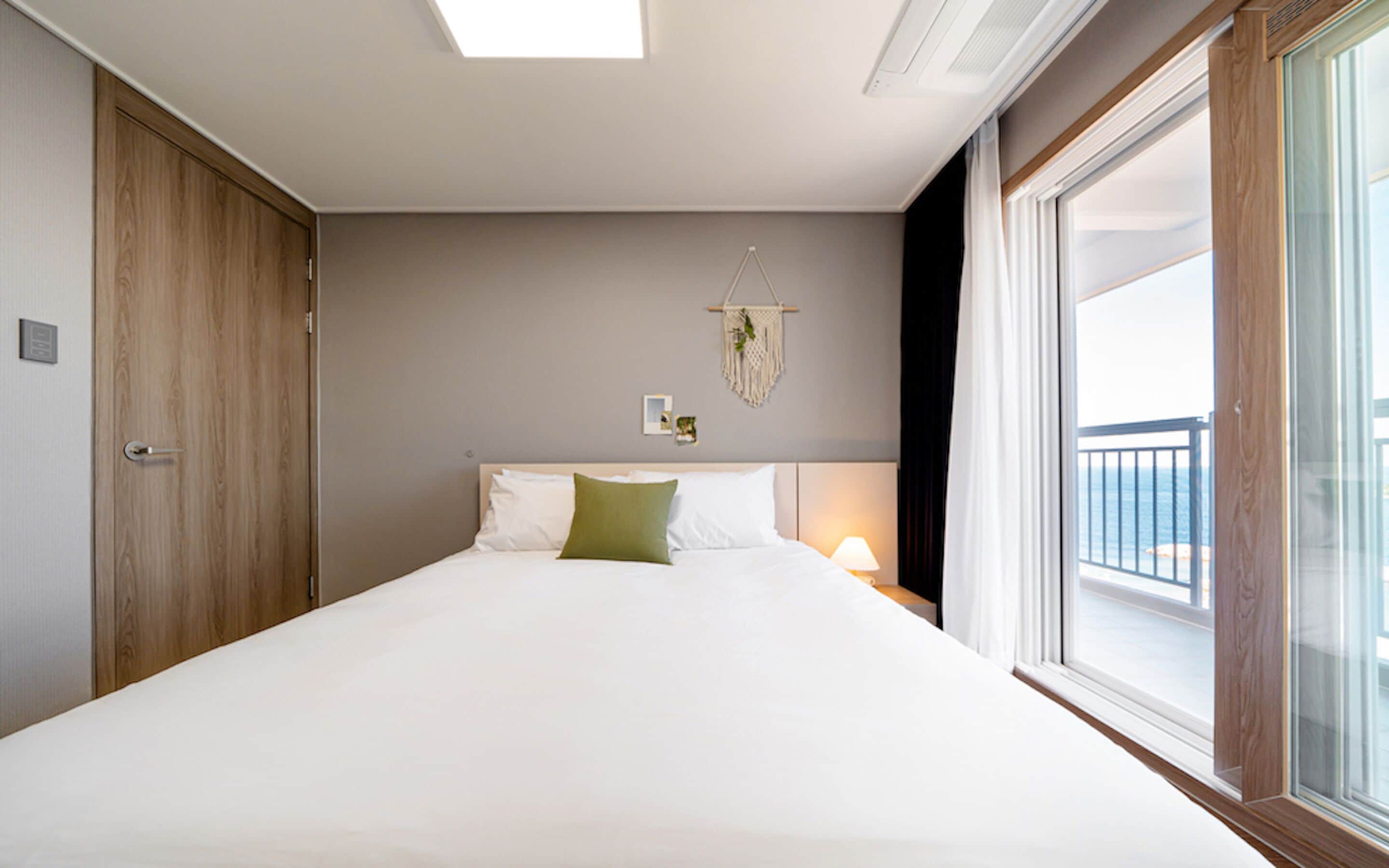 Une chambre avec un grand lit, une porte en bois, une décoration murale en macramé et une fenêtre donnant sur la mer.