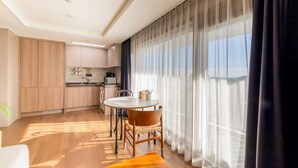 A modern kitchen with wooden cabinets, a dining area with a round table and chairs, and a large window with sheer curtains.