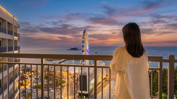 A balcony with a view of a brightly lit Ferris wheel and the sea at dusk.