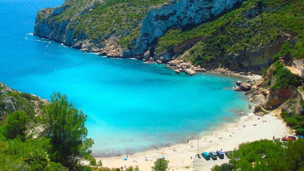 Paesaggio costiero con spiaggia, scogliere rocciose e acque blu cristalline.