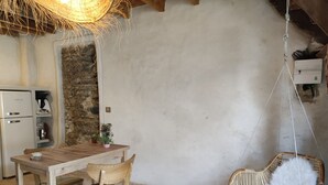 Un comedor rústico con una mesa y sillas de madera, un sillón colgante con cojines blancos, una pared de piedra y una kitchenette con cafetera.