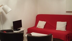 Una stanza piccola con un divano rosso, due cuscini bianchi, una TV nera su un mobile bianco e una lampada bianca.