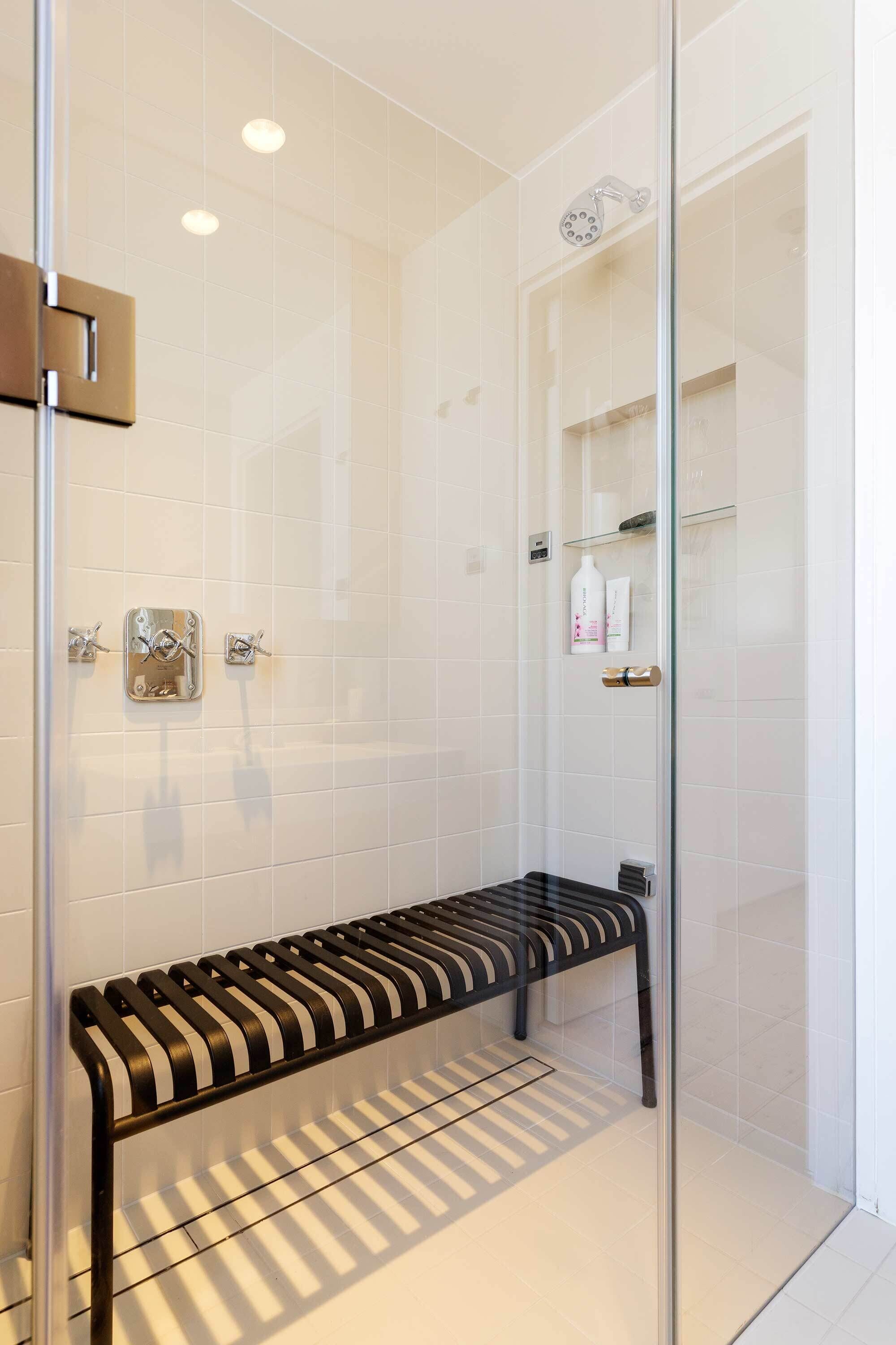 A glass-enclosed shower with a bench, white tiled walls, and a shelf with toiletries.