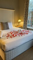 Una cama bien hecha con edredón blanco y almohadas, una lámpara de noche y una ventana con vistas a edificios y árboles.