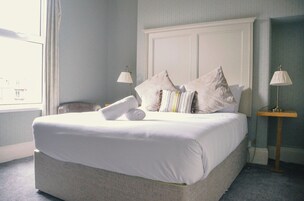 Una cama bien hecha con sábanas y almohadas blancas, una toalla enrollada y dos lámparas de noche.