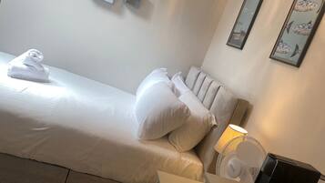 Habitación de hotel con cama, almohadas, mesita de noche con lámpara, ventilador y bandeja con tazas y vaso.