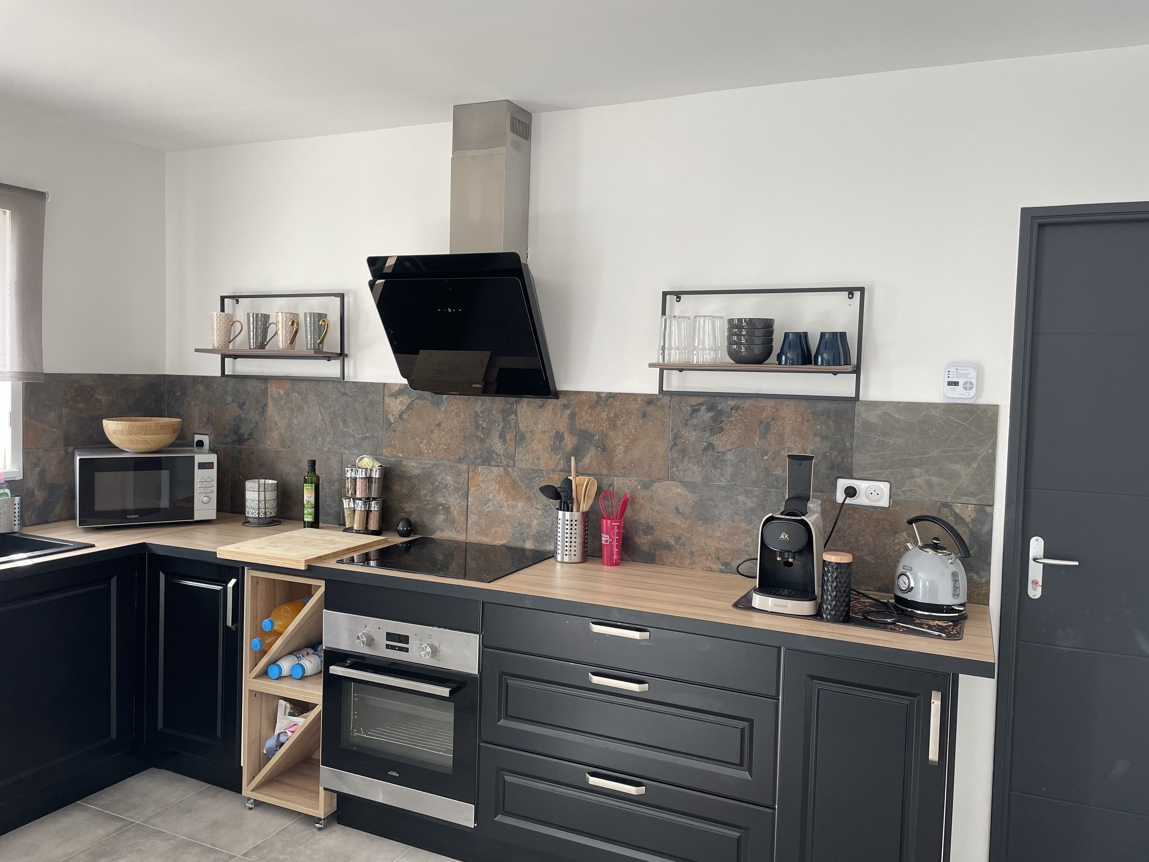 A modern kitchen with a microwave, oven, and coffee maker. There are shelves with cups and a kettle.