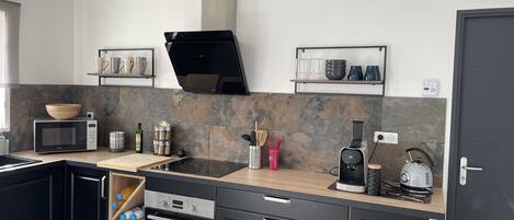 A modern kitchen with a microwave, oven, and coffee maker. There are shelves with cups and a kettle.