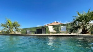 A swimming pool with lounge chairs and a covered area, surrounded by palm trees and a building with a tiled roof. - House Bungalow Private Pool Comfort & Relaxation (Saint-Gilles)