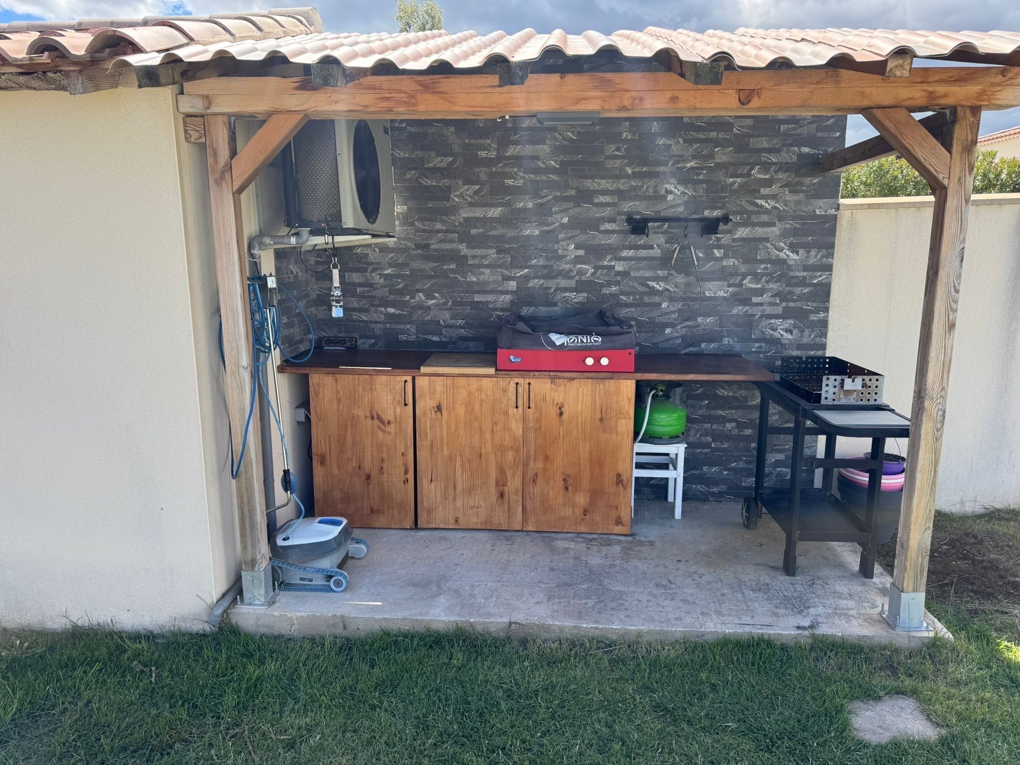 Ett litet utomhuskök med en stenmur, träskåp, en grill och ett bord med en gasolflaska.