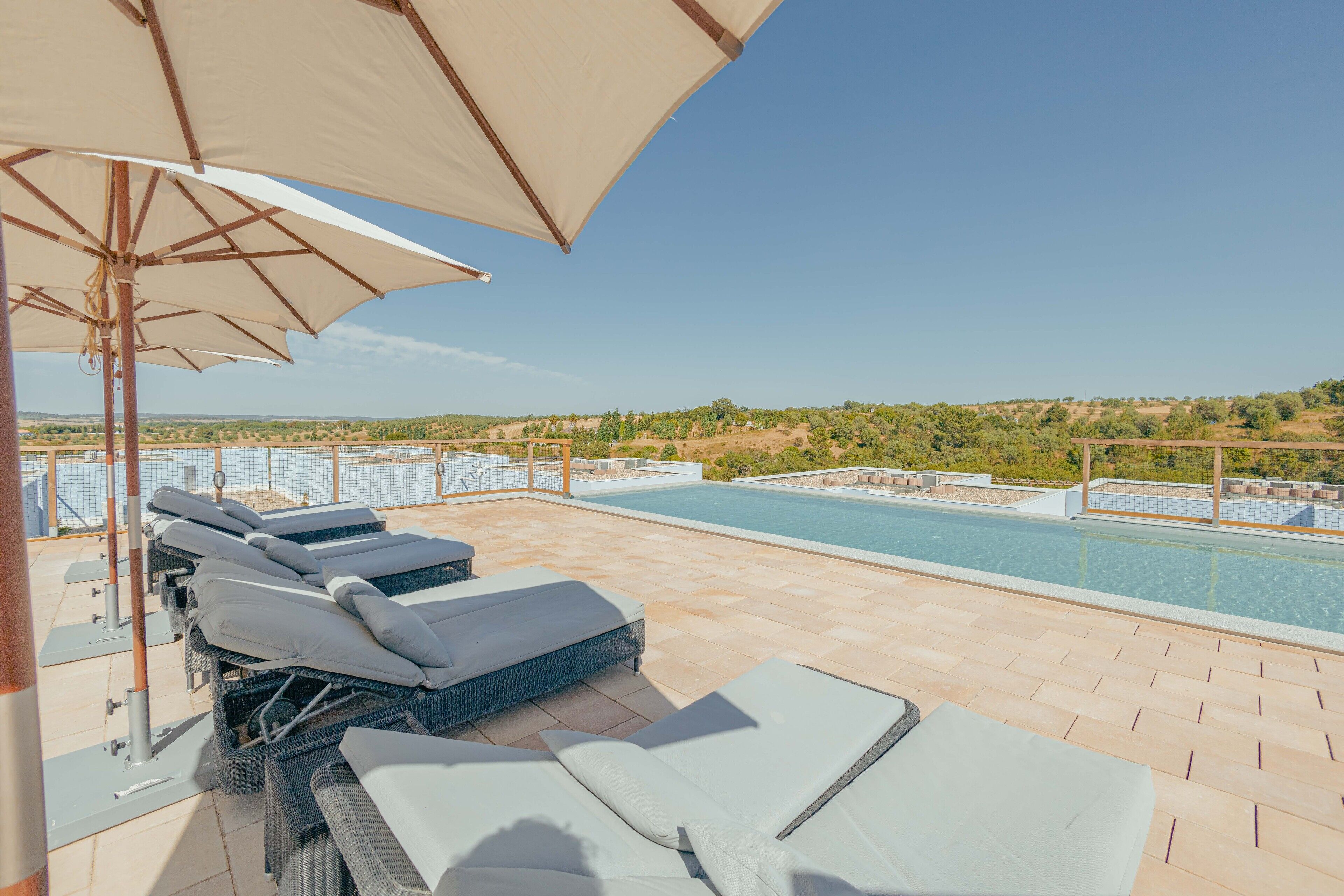 Zona de piscina en la azotea con sillas de descanso y sombrillas, con vistas a un paisaje ajardinado.