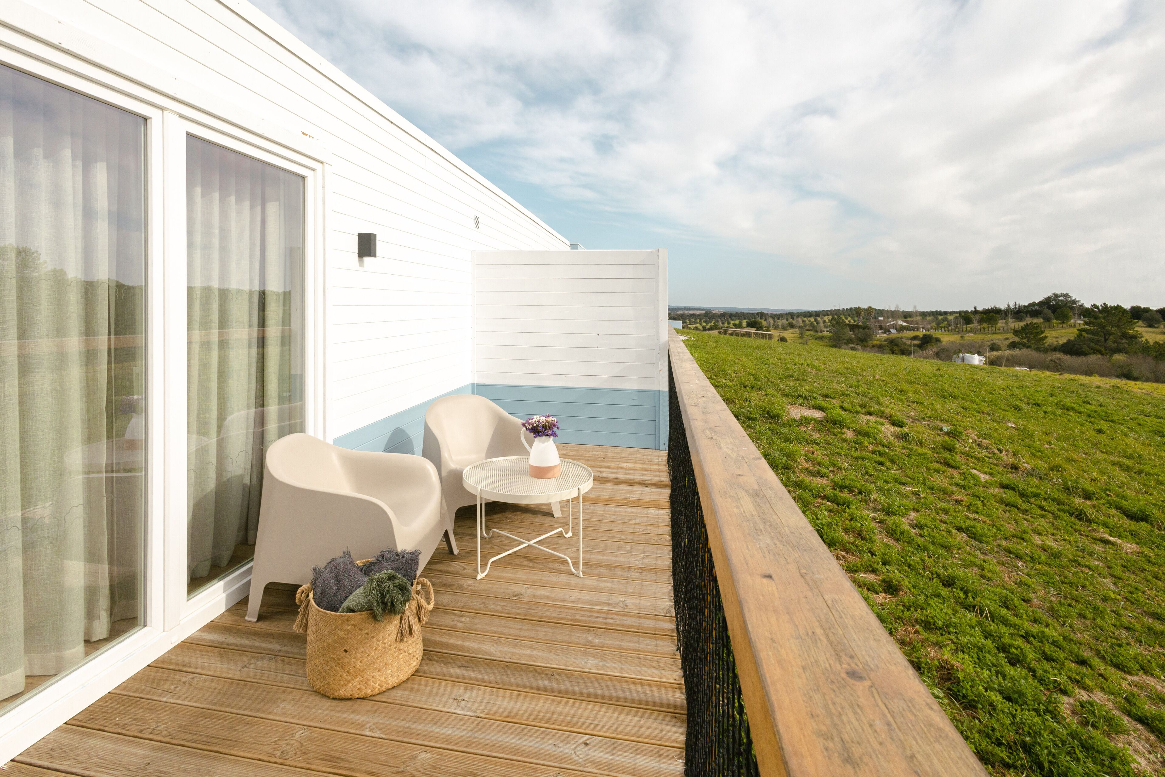 Una terraza de madera con sillas blancas y una pequeña mesa, con vistas a un paisaje cubierto de césped.