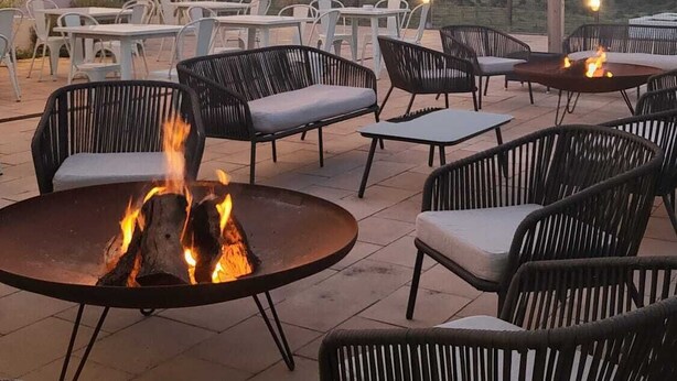 Een terras buiten met tafels en stoelen, een vuurkorf en lichtslinger tegen een achtergrond van een heldere lucht en een verafgelegen landschap.