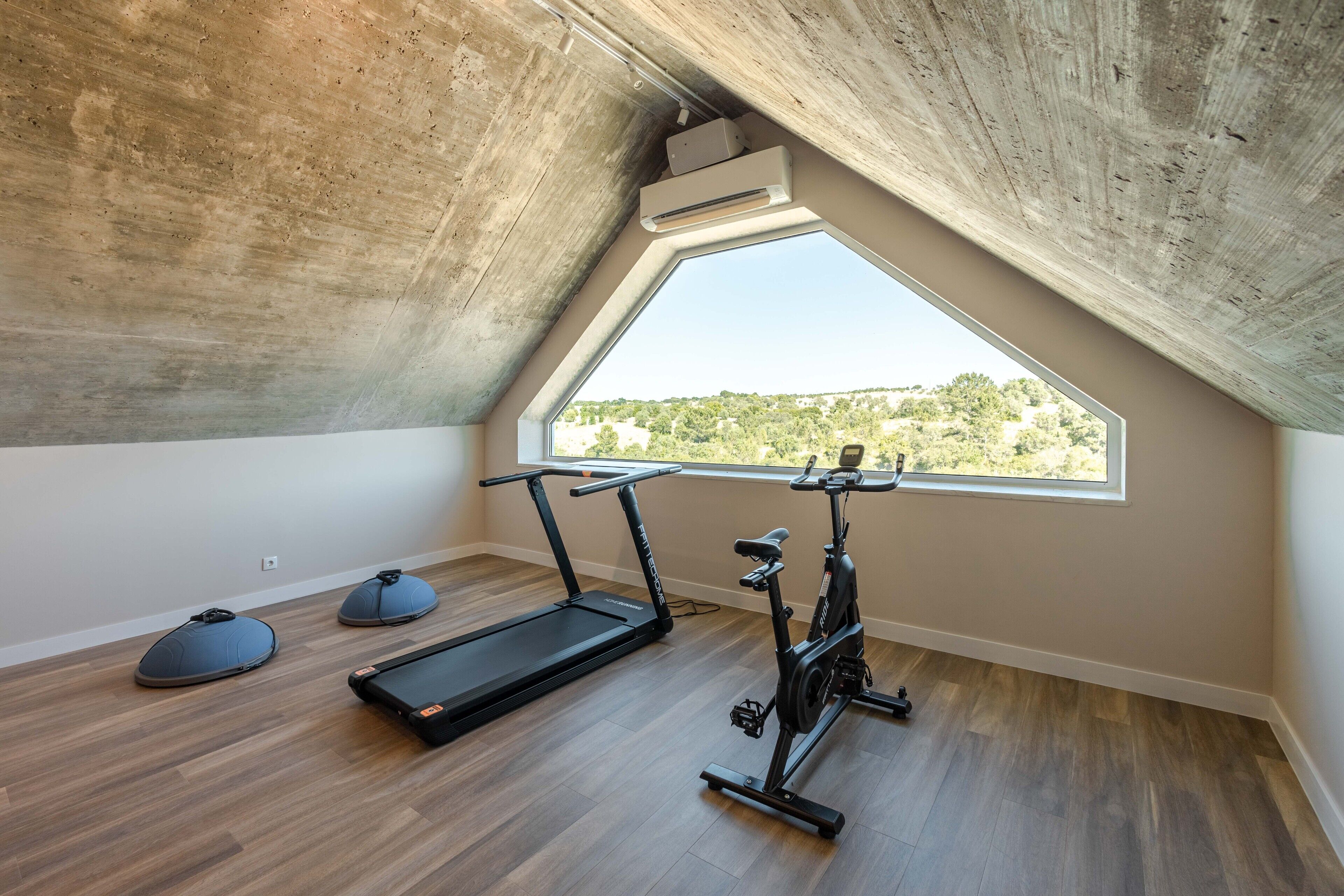 Una habitación pequeña con techo inclinado, un ventanal grande, una cinta de correr y una máquina elíptica.