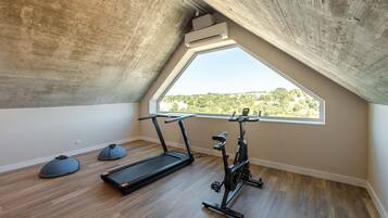 Una habitación pequeña con techo inclinado, un ventanal grande, una cinta de correr y una máquina elíptica.
