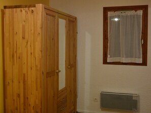 A wooden wardrobe with a mirror, a window with a curtain, and a wall-mounted air vent.