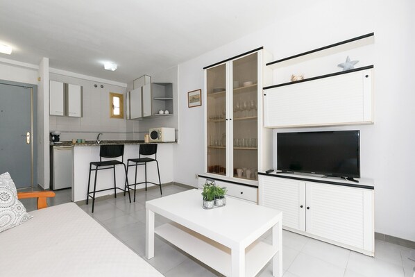 A modern kitchen with white cabinets, a dining area with a white table and two chairs, and a TV on a white cabinet.