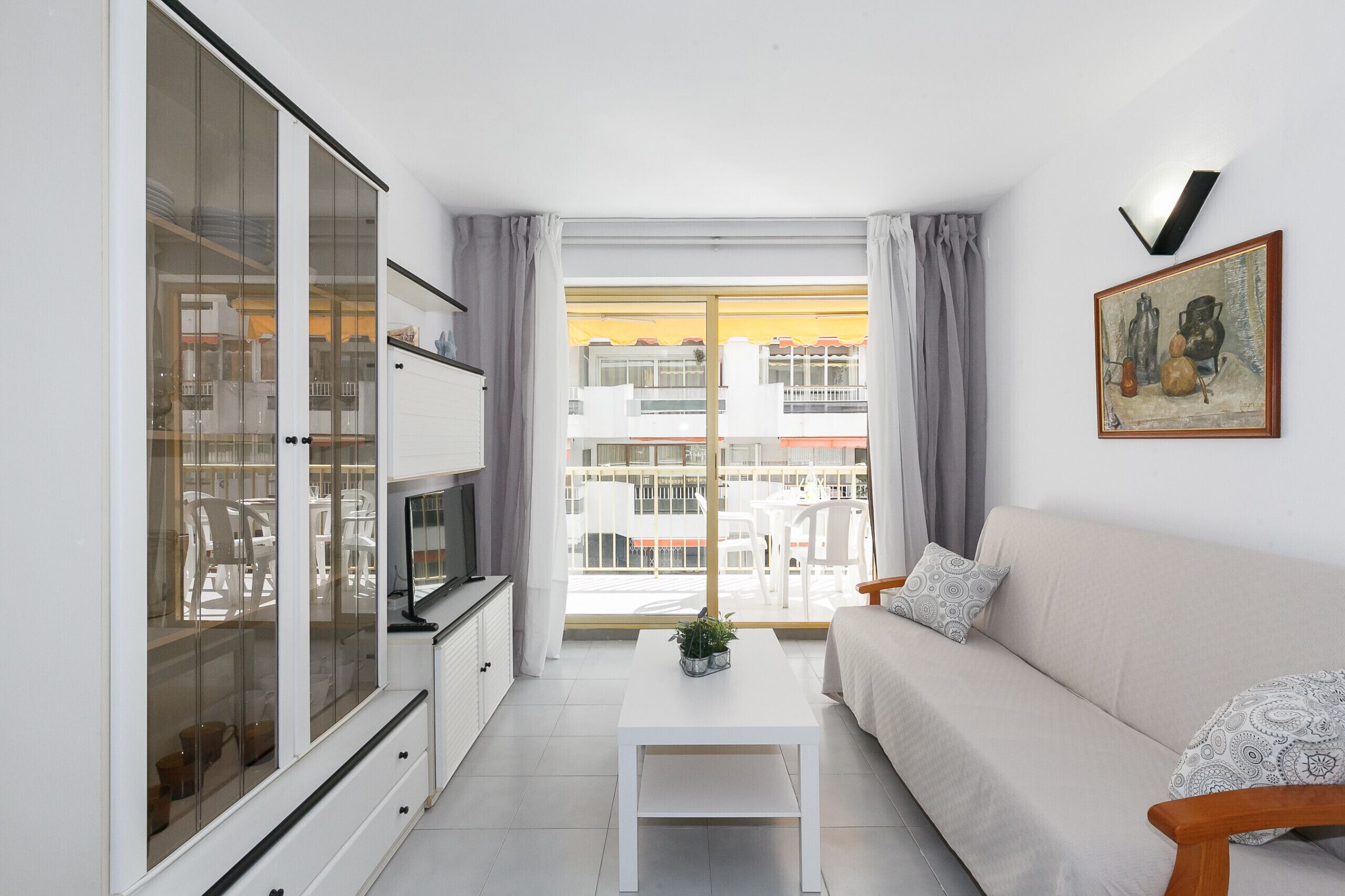 A modern living room with a sofa, a coffee table, and a TV. There is a large window with curtains, and a balcony is visible outside.