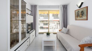 Un salon moderne avec un canapé, une table basse et un téléviseur. Une grande fenêtre avec des rideaux est présente, et on peut apercevoir un balcon à l’extérieur.