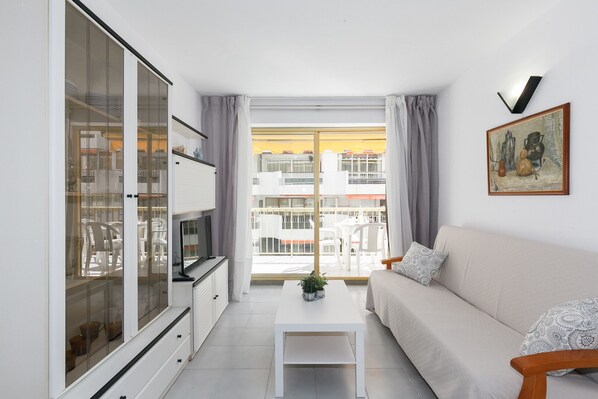 A modern living room with a sofa, a coffee table, and a TV. There is a large window with curtains, and a balcony is visible outside.
