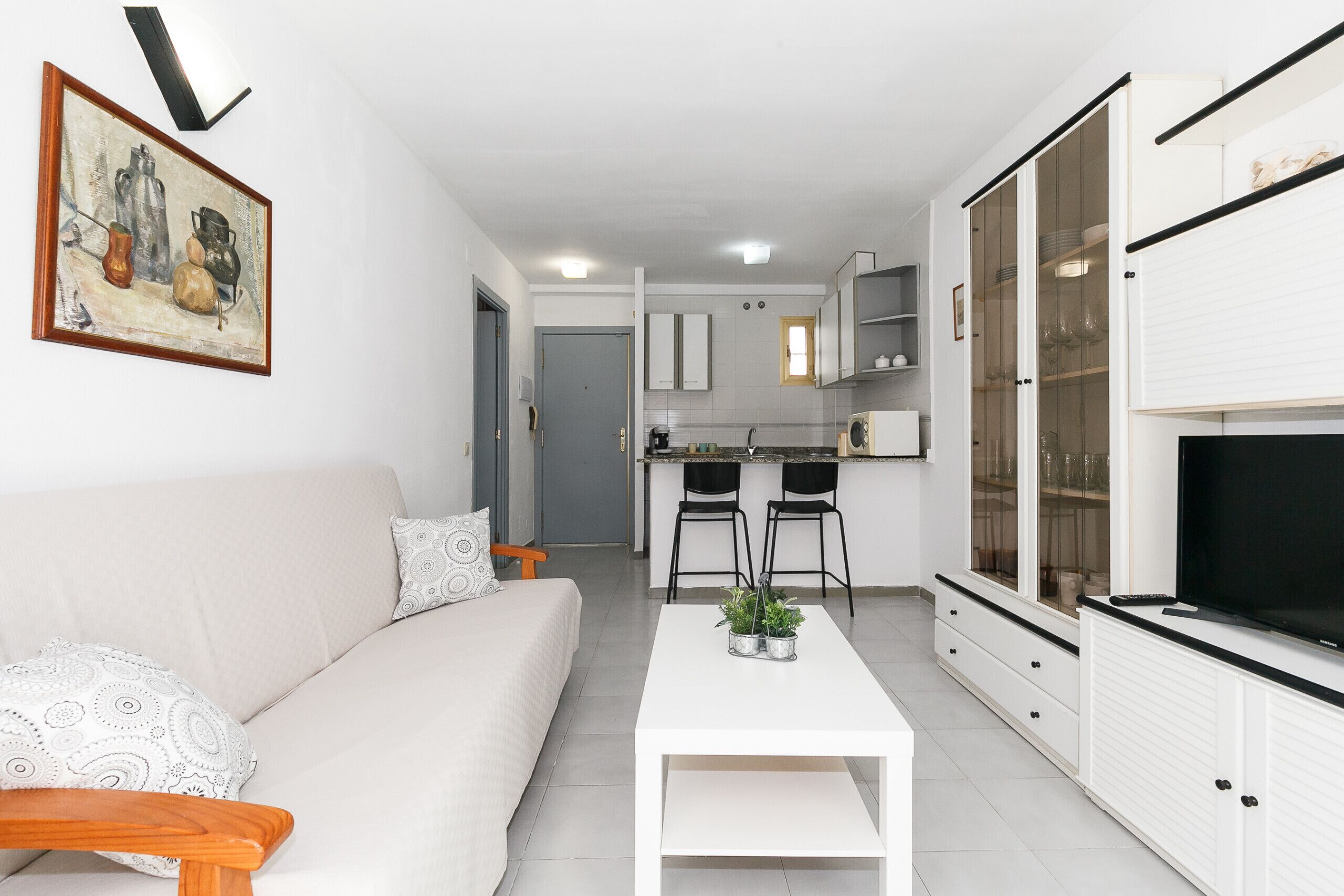 Una sala de estar moderna con sofá, mesa de centro y televisor. Cuenta con una zona de cocina con armarios y un espacio para comer.