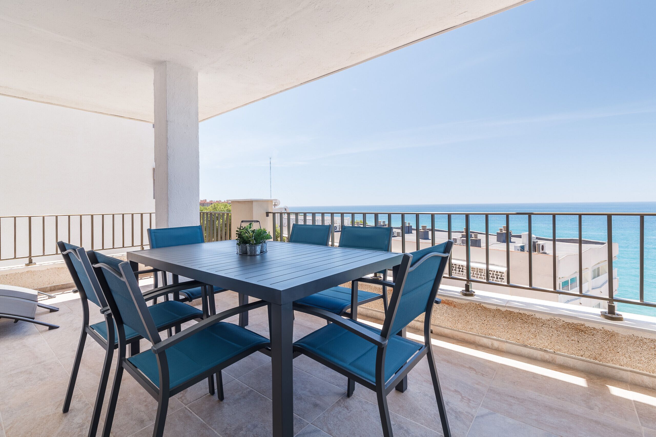 A balcony with a table and chairs, overlooking a beach and the sea.