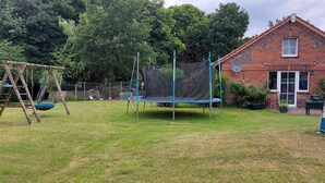 Een speelplaats met een glijbaan, schommel en trampoline, omringd door een bakstenen gebouw en bomen.